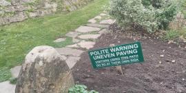 A stone garden path passes before a high stone wall. Near a large decorative rock and in front of a silvery bush, in a wide patch of fresh dirt, a green sign on a low wooden post reads in white capital letters: Polite Warning: Uneven Paving. Visitors using this path do so at their own risk. (That last sentence is not as large print, but is underlined.)