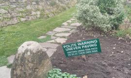 A stone garden path passes before a high stone wall.  Near a large decorative rock and in front of a silvery bush, in a wide patch of fresh dirt, a green sign on a low wooden post reads in white capital letters: Polite Warning: Uneven Paving.  Visitors using this path do so at their own risk.  (That last sentence is not as large print, but is underlined.)